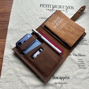 May include: A brown leather organizer wallet featuring a snap closure, a dedicated slot for a smartphone, a pocket for cards, a pen loop with a white pen, and a wooden-covered Field Notes notebook secured inside the leather fold.