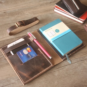 Puede incluir: Una funda de cuero marrón para cuaderno con un bolsillo para un teléfono inteligente y una tarjeta de crédito, un portalápices con un bolígrafo rosa y un cuaderno Moleskine azul. La funda tiene costuras visibles y una correa de cuero separada.