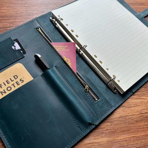 May include: A dark teal leather planner organizer featuring a six-ring metal binder with lined paper, a zippered pocket, a pen loop, a card slot, and a pocket containing a Field Notes notebook and a burgundy United Kingdom passport.