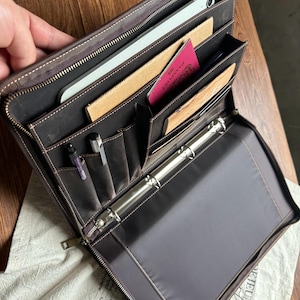 May include: A dark brown leather zippered portfolio organizer featuring a three-ring metal binder mechanism, multiple interior pockets, two pen loops, a tablet sleeve, and slots for a passport and documents, secured with a full-length metal zipper.