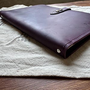 May include: A deep purple leather notebook cover featuring a strap closure with two metallic snap buttons. The cover has visible stitching along the edges and the text "Passion Project" embossed on the front right corner of the leather surface.