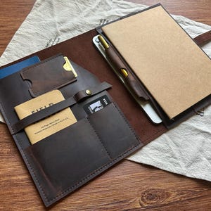 May include: A dark brown leather organiser folio featuring multiple interior pockets for cards and documents, a strap with a brass snap closure, a tan Field Notes memo book, and a notebook with a gold-accented pen held in a leather loop.
