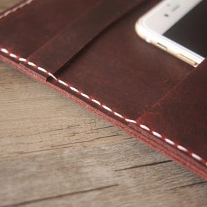 Puede incluir: Una cartera de cuero marrón oscuro con costuras blancas en contraste a lo largo de los bordes. El interior incluye una ranura dedicada que sostiene un teléfono inteligente blanco y un bolsillo pequeño con una tarjeta de flores rojas y blancas.