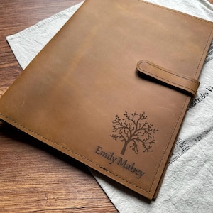 Può includere: Una cartella in pelle color cuoio di forma rettangolare con chiusura a cinturino in pelle abbinata. La copertina anteriore presenta un disegno di un albero inciso al laser e il testo Emily Mabey. La pelle presenta cuciture visibili sui bordi.