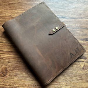 Peut inclure: Une couverture de carnet en cuir marron avec une finition mate lisse et une texture grainée naturelle visible. La couverture comprend une sangle fixée par deux boutons-pression en métal couleur laiton et les initiales A.H. gravées dans le coin.