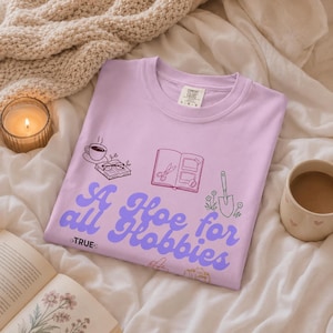 May include: A light purple t-shirt featuring the text A Hoe for all Hobbies in a bold, wavy font. The graphic includes line art illustrations of a coffee cup, an open book with glasses, a scrapbook, and a garden trowel with small flowers.