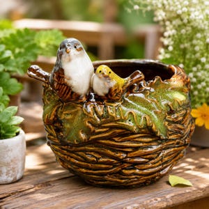 May include: A ceramic planter shaped like a bird nest with a glossy brown finish and green leaf accents. Two small ceramic birds, one larger with white and brown feathers and one smaller with yellow and brown feathers, are perched on the rim.