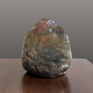 May include: A hand-formed ceramic sculpture with an irregular, rounded shape. The surface features a mottled glaze with earthy tones of deep brown, charcoal black, and muted ochre, displaying a textured, uneven finish with visible kiln-fired marks.