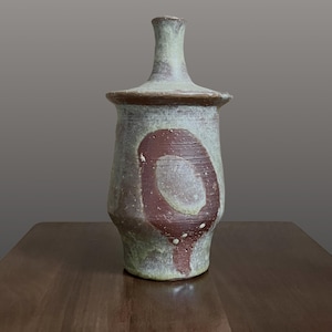 May include: A ceramic bottle with a narrow neck and a wide, flared rim. The body features a textured, mottled light green and grey glaze with a prominent, dark reddish-brown circular pattern on the front. The surface has a visible, rough finish.