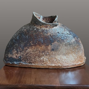 May include: A low, wide ceramic vessel with a rough, textured surface in shades of dark brown, charcoal, and earthy tan. The object features a prominent, uneven central opening with a jagged rim, showcasing a weathered, kiln-fired appearance.