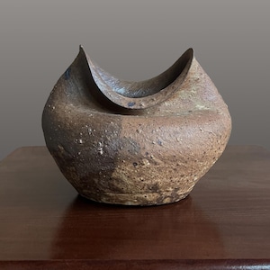 May include: A hand-formed ceramic vessel with a textured, mottled brown and tan surface. The bowl features an asymmetrical, curved rim that dips in the center, creating two distinct points. The exterior has a rough, granular finish.