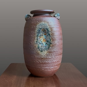 May include: A cylindrical ceramic vase with a textured, reddish-brown exterior featuring horizontal ridges. The center of the vase displays an oval-shaped glaze patch in shades of blue, green, and orange. It has two small, protruding handles.