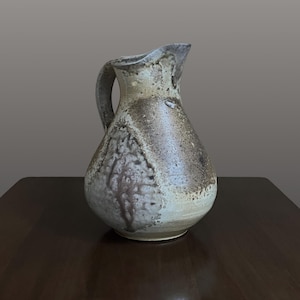 May include: A ceramic pitcher with a bulbous base and a tapered neck, featuring a textured, mottled glaze in shades of cream, beige, and dark brown. The surface displays a rough, earthy finish with visible variations in color and a curved handle.