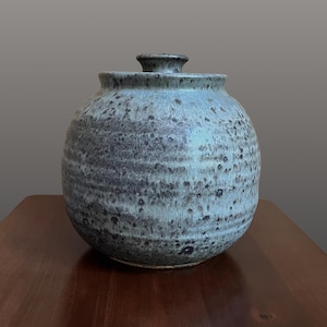 May include: A round ceramic jar with a lid, featuring a mottled glaze in shades of blue, gray, and black. The surface has a textured appearance with horizontal ridges circling the body, and a small, knob-like handle sits atop the center of the lid.