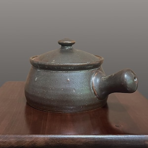 May include: A dark brown, round ceramic pot with a single side handle and a matching lid. The pot features a textured, mottled glaze with subtle variations in tone, a wide base, and a small knob on the center of the lid for easy removal.