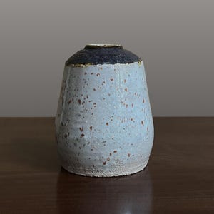 May include: A small ceramic vase with a tapered cylindrical shape. The body features a light blue glaze with scattered brown speckles, while the narrow neck and rim are finished in a dark, textured black glaze with a rough, uneven surface.