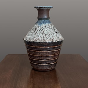 May include: A ceramic vase with a narrow neck and a wide, tapered body. The upper section features a textured, white cross-hatched pattern over a dark brown base, while the lower section displays horizontal ridges in a deep, earthy brown glaze.