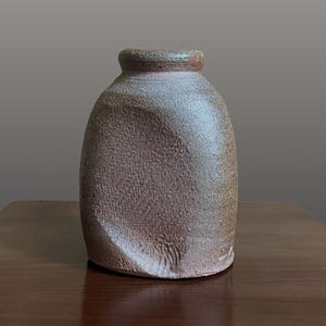 May include: A small, hand-thrown ceramic vase with a textured, earthy brown surface. The vessel features a rounded body with a distinct, indented side panel and a thick, slightly flared rim. The clay has a matte, granular finish throughout.
