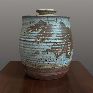 May include: A cylindrical ceramic canister with a lid, featuring a textured surface with horizontal ridges. The glaze is a mix of light blue, cream, and dark brown, with a solid dark brown base and a speckled finish throughout the body.