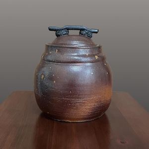 May include: A round, dark brown ceramic jar with a textured, mottled surface and a matching lid. The lid features a distinct, horizontal black handle with a rough, organic texture. The jar has a wide, bulbous body and a smooth, glazed finish.