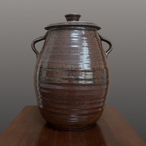 May include: A large, bulbous, dark brown ceramic jar with a glossy, textured finish. The vessel features two curved side handles, a matching lid with a small knob handle, and horizontal ridges encircling the body of the container.