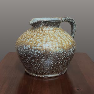 May include: A ceramic pitcher with a rounded body and a single handle. The surface features a mottled glaze with a dark brown base color transitioning to a lighter, textured tan and white speckled pattern near the top rim and spout area.