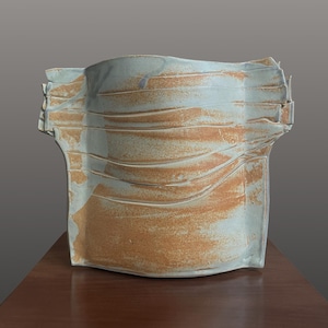 May include: A hand-built ceramic vessel with a wide, flattened rectangular form. The surface features a blend of pale blue-grey glaze and exposed, textured terracotta-colored clay, characterized by horizontal ridges and folded edges.