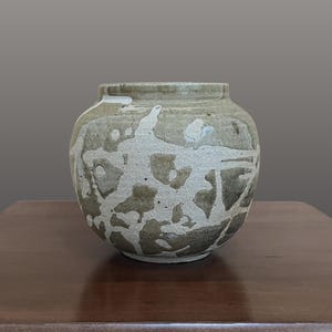May include: A round ceramic vase with a wide opening and a short neck. The surface features a textured, mottled pattern of earthy beige and dark brown tones, created by overlapping layers of glaze that form irregular, organic shapes across the body.