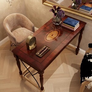 May include: A rectangular mahogany-toned wooden desk with a central oval inlay, four turned legs, and a cross-brace support. The desk surface holds a glass vase with purple flowers, a stack of three books, a gold-toned clock, and a tray with two pens.