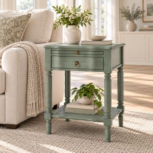 May include: A sage green wooden end table featuring a single drawer with a round brass pull, a pull-out tray with a small brass knob, and a lower shelf. The table stands on four turned legs and has a smooth, painted finish with subtle distressing.