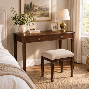 May include: A dark wood writing desk with two drawers featuring round brass pulls, fluted legs, and a matching upholstered stool with a beige seat. The desk surface holds a white ceramic vase with greenery, stacked books, and a brass table lamp.