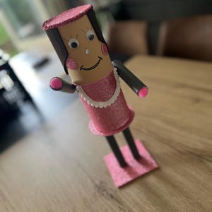 May include: A handcrafted figure made from a cylindrical tube with a tan face, googly eyes, and a drawn smile. It features a pink glittery hat, a pink glittery dress with a white scalloped collar, black arms with pink tips, and black legs.