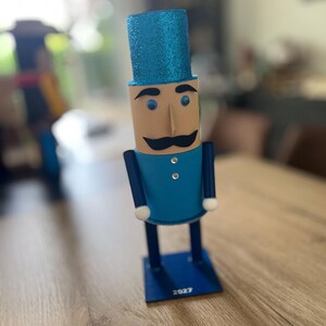 May include: A nutcracker figurine featuring a bright blue glitter top hat, a tan face with a black mustache, and a blue uniform jacket with two small silver buttons. The figure stands on a blue square base with the year 2027 printed in white.
