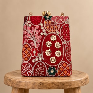 Op de afbeelding: Een rechthoekige rode clutch met ingewikkeld borduurwerk in bloem- en geometrische patronen met roze, oranje en groene draad. Het oppervlak is versierd met goudkleurige lovertjes en een metalen gouden sluiting aan de bovenkant in het midden.