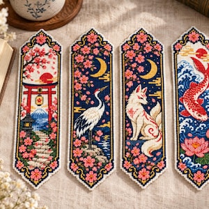 May include: Four rectangular fabric bookmarks featuring cross-stitch embroidery. Designs include a red torii gate with Mount Fuji, a white crane, a multi-tailed fox, and a red koi fish, all surrounded by pink cherry blossoms and dark blue backgrounds.