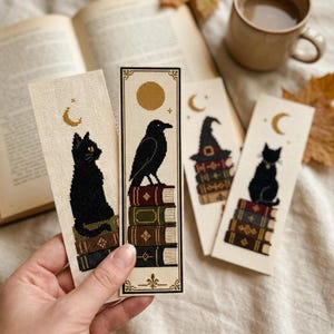 Peut inclure: Quatre marque-pages rectangulaires en tissu ornés de broderies au point de croix. Les motifs représentent des chats noirs, un corbeau noir et un chapeau de sorcière, posés sur des piles de livres colorés. Chaque marque-page présente des motifs célestes.