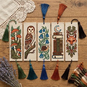 May include: Five rectangular fabric bookmarks featuring cross-stitch embroidery designs of mushrooms, an owl, bluebells with a snail, a stack of books with a crescent moon, and a fox face, each finished with a matching colored thread tassel.