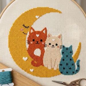 Peut inclure: Un projet de point de croix dans un tambour en bois représentant un croissant de lune jaune avec un visage, un chat orange, un chat crème et un chat bleu sarcelle à pois noirs. Des petits cœurs blancs sont dispersés autour des chats.