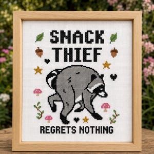 May include: A framed cross-stitch embroidery featuring a gray raccoon in the center with the black text SNACK THIEF above and REGRETS NOTHING below. The design includes small colorful motifs of green leaves, brown acorns, gold stars, black hearts, and pink mushrooms.