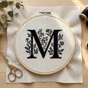 Puede incluir: Un bastidor de madera que contiene una tela Aida de color blanco roto con una letra M bordada en punto de cruz negro y rodeada de motivos florales. Junto al bastidor hay unas tijeras de bordar doradas y varios carretes de hilo DMC negro.