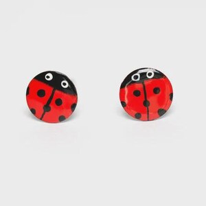 Pode incluir: Um par de brincos circulares com design de joaninha. Cada brinco tem um fundo vermelho brilhante com pintas pretas e uma seção de cabeça preta contendo dois olhos circulares brancos. A superfície é lisa e apresenta um acabamento brilhante.