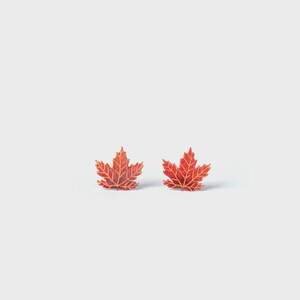 Peut inclure: Une paire de boucles d'oreilles à clous en forme de feuilles d'érable. Chaque boucle présente une finition rouge vif avec des nervures dorées en relief détaillées qui suivent la structure naturelle de la feuille, offrant un aspect texturé.
