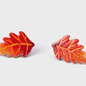 May include: A pair of stud earrings shaped like oak leaves. The leaves feature a vibrant gradient of red, orange, and yellow enamel with raised, textured veins in a lighter yellow tone, creating a detailed and colorful autumn-inspired design.