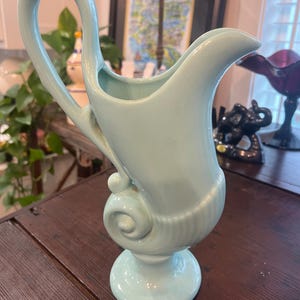 May include: A light blue ceramic pitcher featuring a tall, curved handle and a flared pouring spout. The base of the vessel is circular, supporting a body with a prominent spiral scroll design on the side and vertical ribbed detailing.