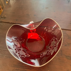 May include: A deep red glass bowl with a wavy, silver-trimmed rim. The bowl features etched floral designs on the interior sides and a small, bright red rectangular tag attached to the center base of the interior.