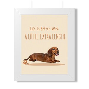 May include: A framed wall art print featuring a brown dachshund lying down on a light beige background. The text reads Life Is Better With A LITTLE EXTRA LENGTH in brown letters. A small brown paw print graphic is located near the dog.