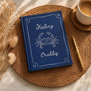 May include: A dark blue hardcover journal featuring a white line-art illustration of a crab in the center, with the text "Feeling Crabby" printed above and below the crab, enclosed in a decorative white border with small heart accents at corners.