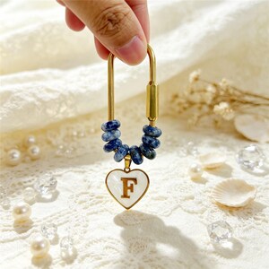 Peut inclure: Un porte-clés mousqueton en forme de U doré, orné de six perles rondes bleu marbré enfilées sur la courbe inférieure, avec un pendentif en forme de cœur blanc suspendu au centre, affichant une lettre F dorée bien visible sur sa surface.