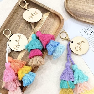 Peut inclure: Trois porte-clés ronds en bois avec des fermoirs en métal doré. Chaque porte-clé présente un nom personnalisé gravé en écriture cursive : Lizbeth, Marleigh et Sulley. Des pompons en tissu coloré à plusieurs niveaux sont attachés.