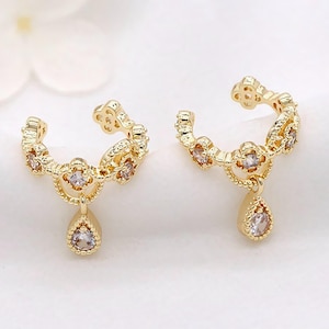 May include: A pair of gold-colored ear cuffs. Each cuff features a floral design with small, clear gemstones and a teardrop-shaped pendant. The earrings are displayed on a white surface.